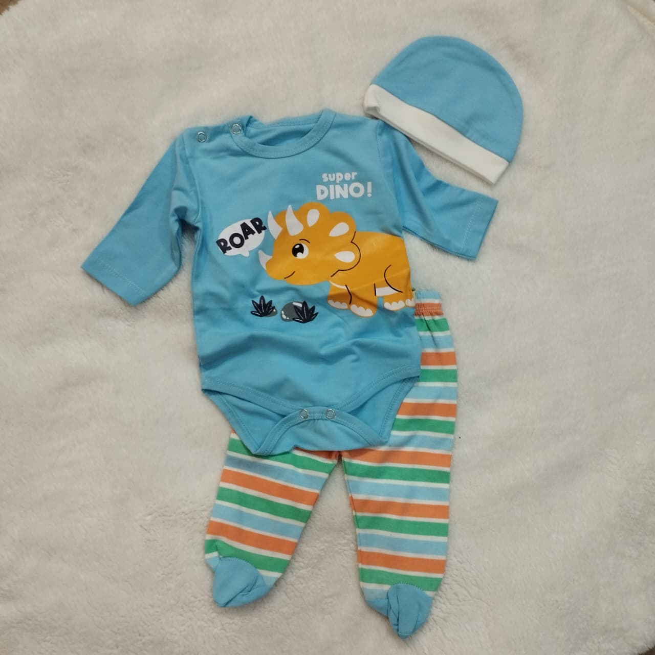 Baby outfit set with blue bodysuit, striped pants, and hat featuring a dinosaur design on a beige blanket.